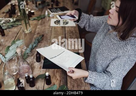 Hochwinkelansicht der mittleren erwachsenen weiblichen Besitzerin, die Broschüre fotografiert Beim Sitzen am Tisch in der Werkstatt Stockfoto