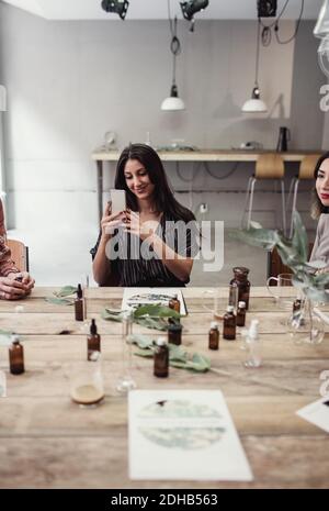 Lächelnde junge Frau, die von Smartphone aus fotografiert, während sie inmitten sitzt Kollegen am Tisch in Parfüm-Werkstatt Stockfoto