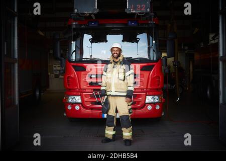 In voller Länge Porträt von lächelnden männlichen Feuerwehrmann vor stehen Des Feuerwehrmotors in der Feuerwache Stockfoto