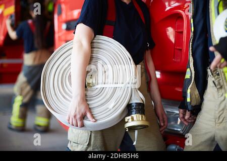 Mittelteil der weiblichen Feuerwehrmann halten zusammengerollt Feuerwehrschlauch während Stehen mit Kollegen an der Feuerwache Stockfoto