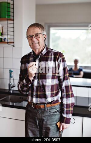 Porträt eines lächelnden älteren Mannes, der auf dem Smartphone durch spricht In-Ear-Kopfhörer, während Sie zu Hause in der Küche stehen Stockfoto