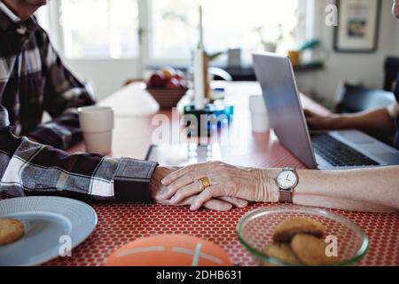 Beschnittenes Bild einer älteren Frau, die die Hände mit dem Mann hält Esstisch zu Hause Stockfoto