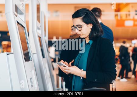 Geschäftsfrau, die am Flughafen ein Mobiltelefon mit einem automatischen Check-in-Automaten nutzt Stockfoto