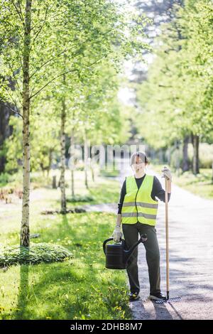 In voller Länge Porträt der lächelnden jungen Frau hält Gießkanne Und Rechen auf Fußweg im Garten Stockfoto