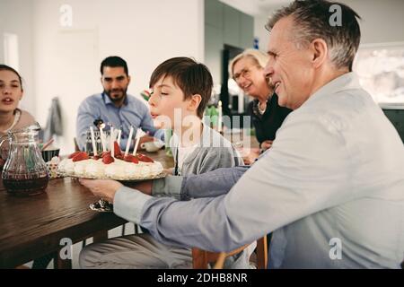 Junge bläst Kerzen auf Geburtstagskuchen, während Sie mit der Familie sitzen Während der Party Stockfoto