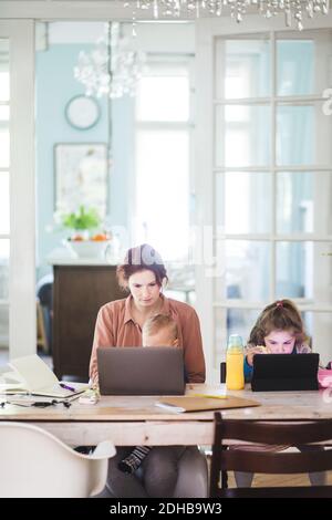 Frau, die am Laptop arbeitet, während sie mit Kindern am Tisch sitzt Stockfoto