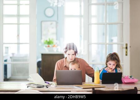 Frau mit mittlerem Erwachsenen, die am Laptop arbeitet, während sie mit Kindern sitzt Zu Hause Stockfoto