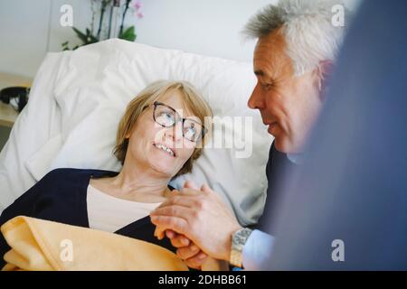 Lächelnde Frau, die auf dem Bett liegend zu einem älteren Partner schaut Auf der Krankenstation Stockfoto