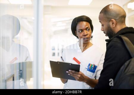 Weiblicher Doktor, der mit männlichem Patienten über Zwischenablage bei beleuchtet diskutiert Korridor im Krankenhaus Stockfoto