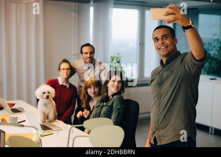 Lächelnder Geschäftsmann nimmt Selfie mit Kollegen bei Hund bei Creative Büro Stockfoto