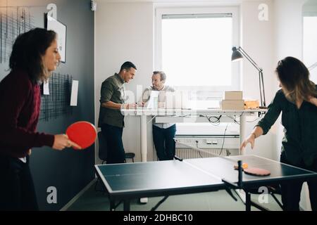 Weibliche Profis spielen Tischtennis, während männliche Kollegen mit Laptop Im Kreativbüro Stockfoto