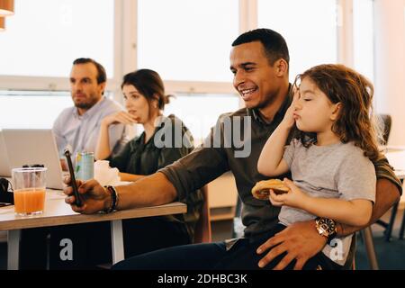 Lächelnder Geschäftsmann nimmt Selfie mit Sohn, während er von Kollegen sitzt Im Kreativbüro Stockfoto