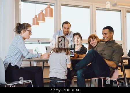 Lächelnder Geschäftsmann, der dem Sohn ein Smartphone zeigt, während er vorsitzt Kollegen im Kreativbüro Stockfoto