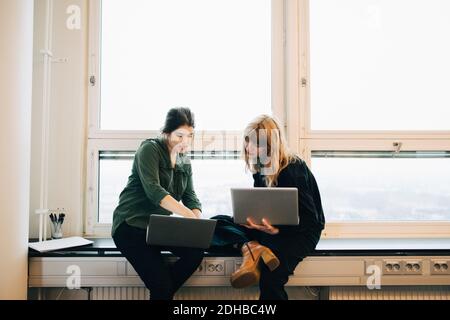 Selbstbewusste Kolleginnen diskutieren über Laptops, während sie am Fenster sitzen Schweller im Kreativbüro Stockfoto