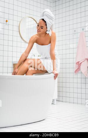 Lächelnde Frau in Handtüchern, die das Bein berührten, während sie im Bad saß Stockfoto
