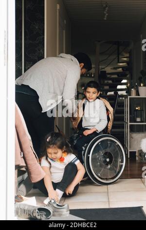 Vater, der Sohn beim Tragen einer Jacke unterstützt, während die Tochter darauf sitzt Boden an der Tür Stockfoto