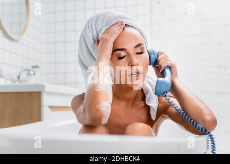 Erschöpfte Frau in Schaum und Handtuch auf dem Kopf reden Telefon beim Baden Stockfoto