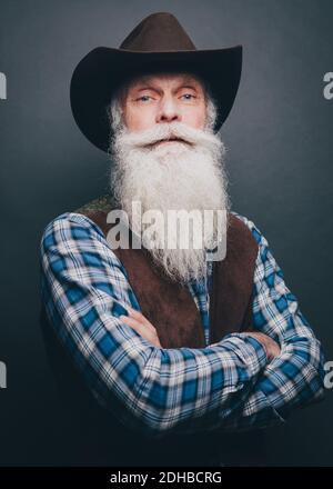 Bärtiger älterer Mann mit Cowboyhut und gekreuzten Armen Grauer Hintergrund Stockfoto