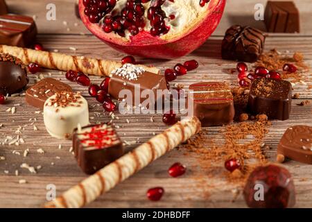 Halb geschnittener Granatapfel mit Samen auf Holztisch verstreut mit Keksstäbchen, verschiedene Pralinen, Kakaopulver und Kokosnussspäne Gattungen bedeckt Stockfoto