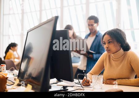 Fokussierte Geschäftsfrau mit Computer, während Mitarbeiter im Hintergrund arbeiten Büro Stockfoto