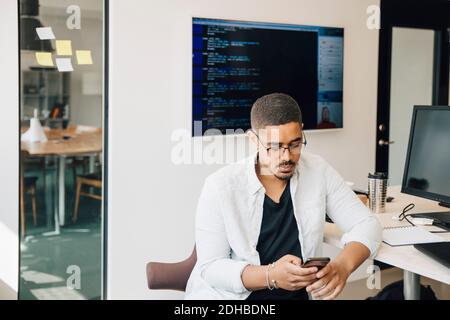 Männlicher Computerhacker, der Smartphone benutzt, während er am Schreibtisch sitzt Im kreativen Büro Stockfoto