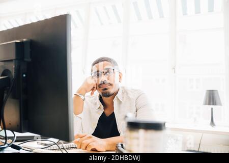 Nachdenklicher Computerprogrammierer, der wegschaut, während er beim Fenster innen sitzt Büro Stockfoto