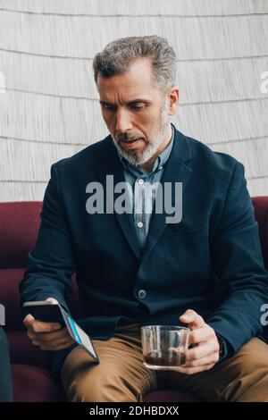 Reifer Geschäftsmann mit Tee, während mit Smartphone auf der Couch Im Büro Stockfoto