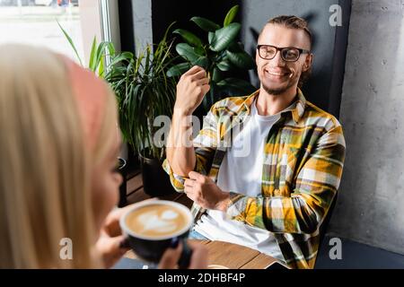 Lächelnder junger Mann in einer Brille, der Frau beim Becherhalter ansieht Von Kaffee auf verschwommenem Vordergrund Stockfoto