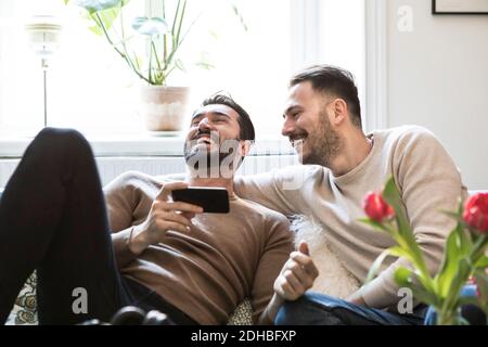 Fröhliches Gay-Paar mit Smartphone sitzt auf dem Sofa gegen Fenster zu Hause Stockfoto