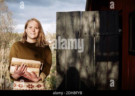 Porträt einer lächelnden jungen Frau, die Feuerholz hält, während sie draußen steht Hütte Stockfoto