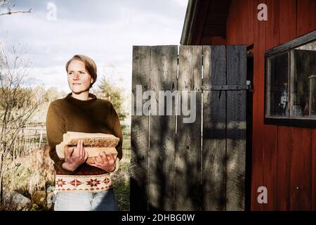 Nachdenkliche junge Frau, die Feuerholz hält, während sie vor der Hütte steht Stockfoto