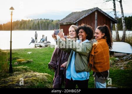 Glückliche Freundinnen nehmen Selfie mit Smartphone im Stehen Auf Grasfeld gegen See bei Sonnenuntergang Stockfoto