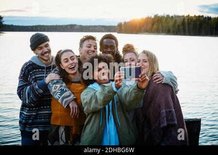 Glückliche männliche und weibliche Freunde, die Selfie mit dem Smartphone machen Während des Sonnenuntergangs gegen den See stehen Stockfoto
