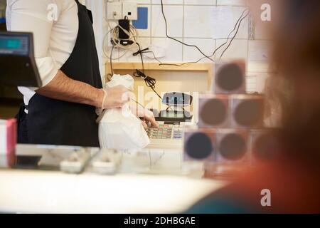 Mittelteil der männlichen Kassierer hält Plastiktüte, während mit Bargeld registrieren Sie sich im Geschäft Stockfoto