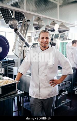 Porträt eines lächelnden männlichen Küchenchefs, der mit der Hand auf der Hüfte steht In der Küche Stockfoto