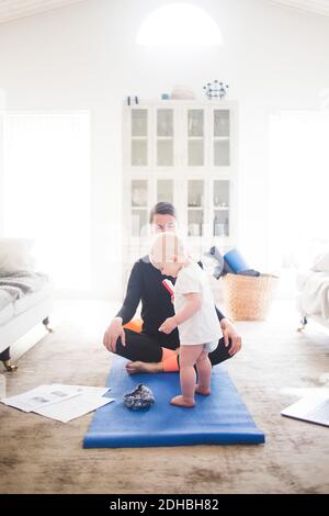 Berufstäbliche Mutter praktiziert Yoga, während Tochter auf Übungsmatte steht Zu Hause Stockfoto