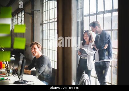 Fröhlicher Geschäftsmann, der am Schreibtisch sitzt, während IT-Profis arbeiten Hintergrund durch Glas gesehen Stockfoto