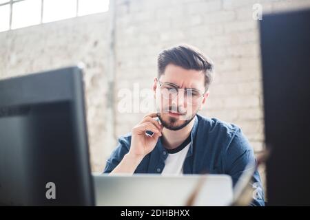 Nachdenkliche junge IT-Experte berühren Bart beim Blick auf Laptop Im Büro Stockfoto
