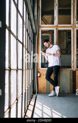 Volle Länge der zuversichtlich männlichen Hipster Hacker mit Smartphone Am Fenster im Kreativbüro Stockfoto