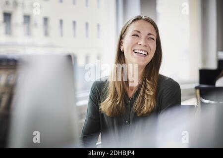 Lächelnde Geschäftsfrau schaut weg, während sie am Fenster im Büro sitzt Stockfoto