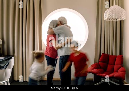 Ältere Paare umarmen, während Enkelkinder im Zimmer spielen Stockfoto