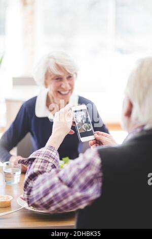 Älterer Mann hält Handy, während er mit einer fröhlichen Frau sitzt Am Tisch im Pflegeheim Stockfoto