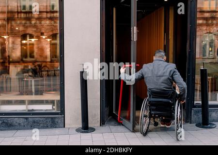 Rückansicht des behinderten älteren Mannes, der auf dem Rollstuhl sitzt, während Im Geschäft betreten Stockfoto