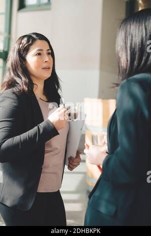 Weibliche Profis, die Kaffee trinken, während sie im Bürokorridor diskutieren Stockfoto