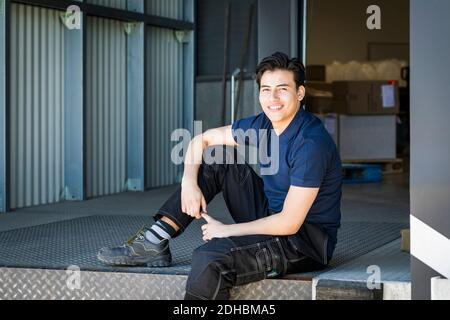 Porträt eines lächelnden jungen männlichen Arbeiters, der am Eingang des Lagerhauses sitzt Stockfoto