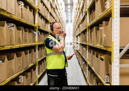 Leitender männlicher Arbeiter mit digitalem Tablet, der Pakete auf sieht Rack während des Gesprächs durch das Headset bei der Distribution Warehou Stockfoto