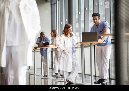 Junge Krankenschwester diskutieren mit mittleren Erwachsenen Arzt über Laptop, während Im Krankenhaus gegen das Fenster stehen Stockfoto