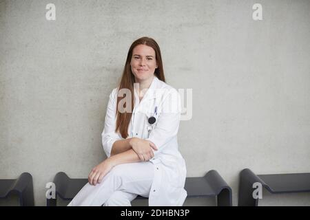 Portrait von lächelnd junge weibliche Brünette Arzt sitzt gegen die Wand Im Krankenhaus Stockfoto