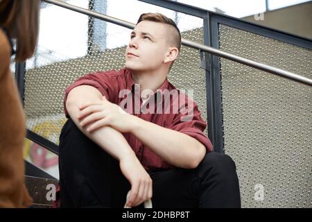 Junger Mann schaut weg, während er auf Stufen gegen Geländer sitzt An der Universität Stockfoto