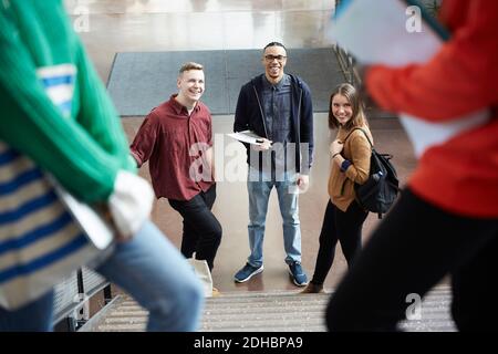 Lächelnde Freunde, die während der Zeit weibliche Studenten auf Stufen betrachten Im Flur stehend Stockfoto